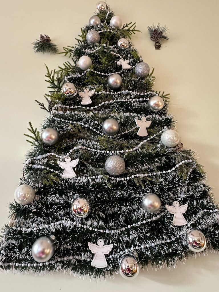 Dekoracja świąteczna: Płaska choinka na ścianie zrobiona z zielonego łańcucha, ozdobiona srebrnymi koralikami, bombkami i białymi, drewnianymi aniołkami. 