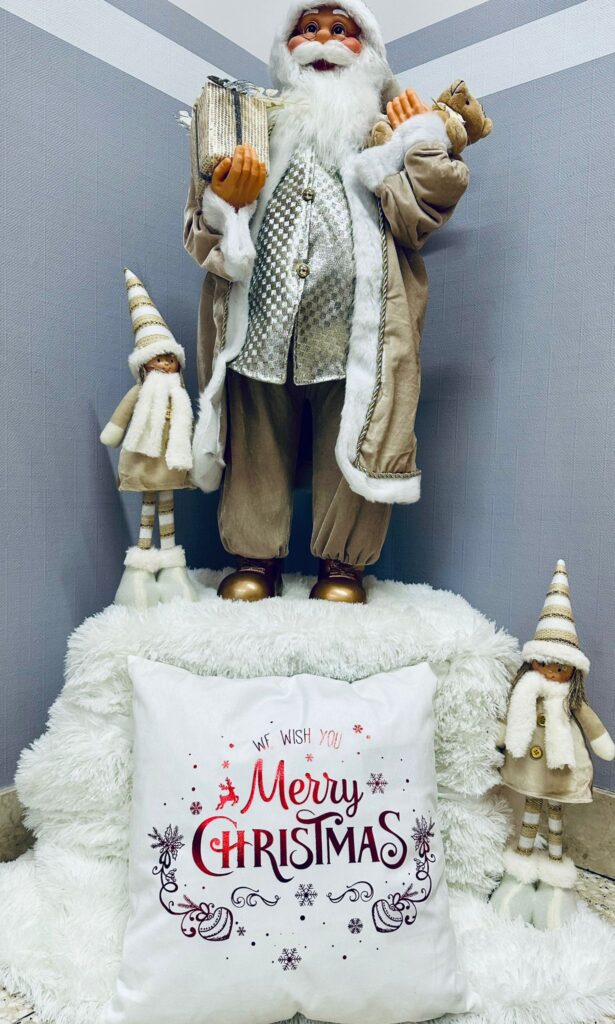 Dekoracja świąteczna: podwyższenie okryte białym, puchowym kocem, przed którym położona jest poduszka z napisem "Merry Christmas", na podwyższeniu stoi duża figura starszego mężczyzny (podobnego do Mikołaja) w srebrno beżowym stroju oraz figurki elfów. 