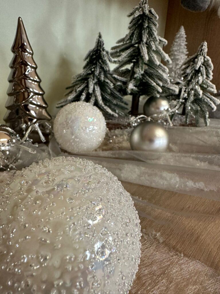 Dekoracja świąteczna: na blacie szafki rozłożony jest biały tiul, na którym ułożone są malutkie choineczki i złote oraz srebrne kule. 