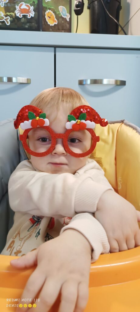 Mały chłopczyk w czerwonych okularach z ozdobami w kształcie mikołajowych czapek nad oprawkami. 