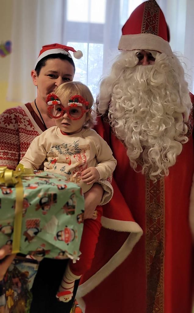 Kobieta z małym dzieckiem na rękach pozują do zdjęcia z mikołajem. Dziecko ma na nosie duże czerwone oprawki okularów ze świątecznymi ozdobami. 