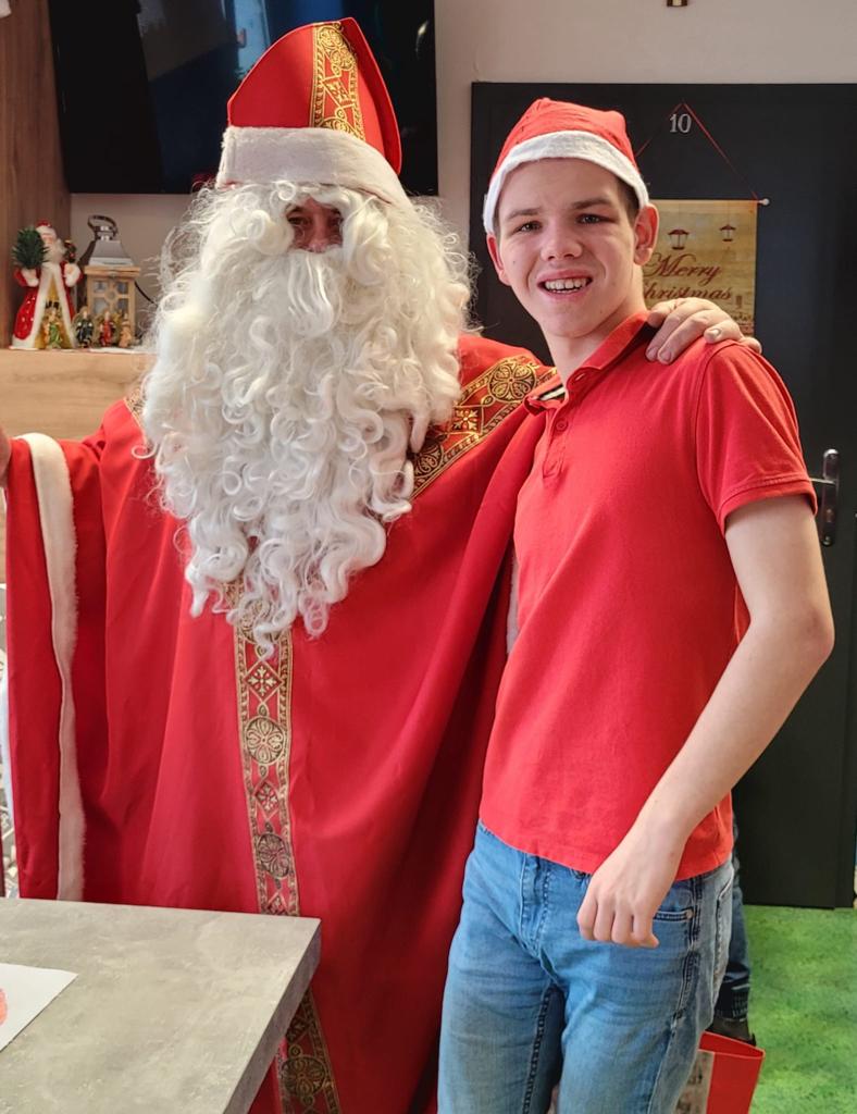 Chłopak w czerwonej koszulce i mikołajkowej czapce pozuje do zdjęcia z Mikołajem. 