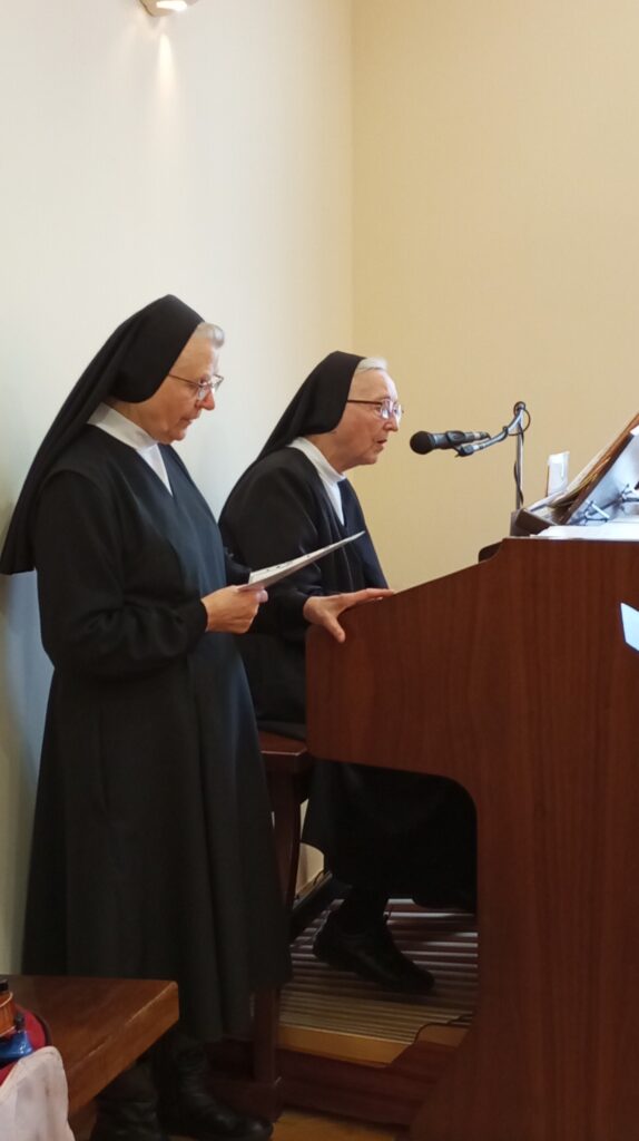 Dwie starsze siostry zakonne śpiewają do mikrofonu. Jedna z nich gra na organach. 