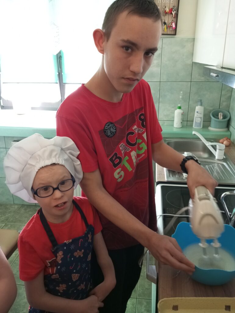Dwoje chłopców w kuchni. Jeden w czapce kucharskiej na głowie i fartuszku uśmiecha się do aparatu. Drugi miksuje coś w misce. 