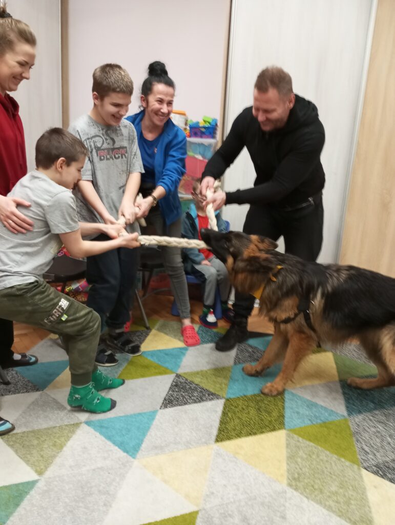 Chłopcy podczas zajęć dogoterapeutycznych - głaskający psa, siłujący się z nim liną, czeszący. 