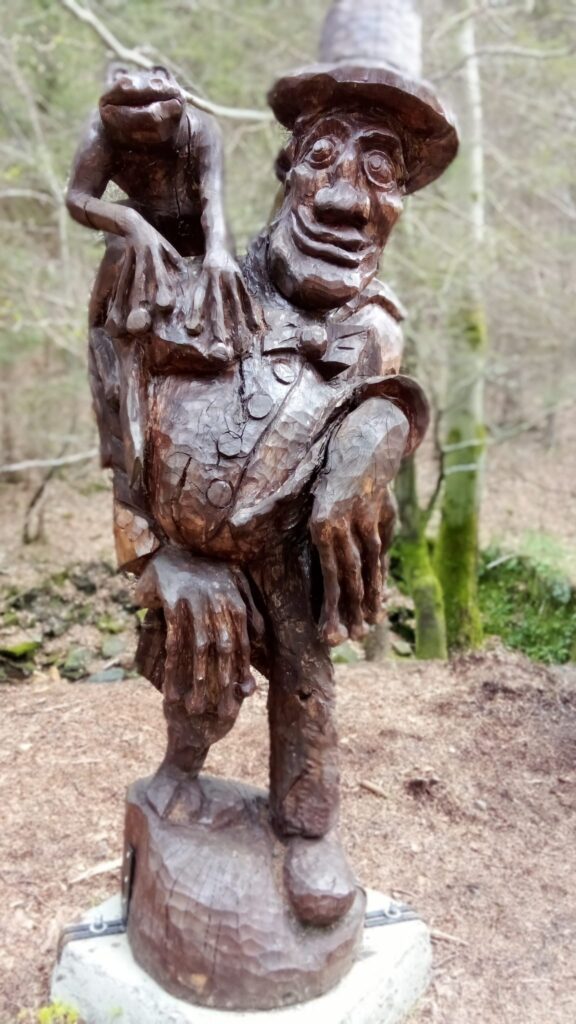 drewniana rzeźba człowieka w muszce i cylindrze na głowie. Na jego ramieniu siedzi żaba